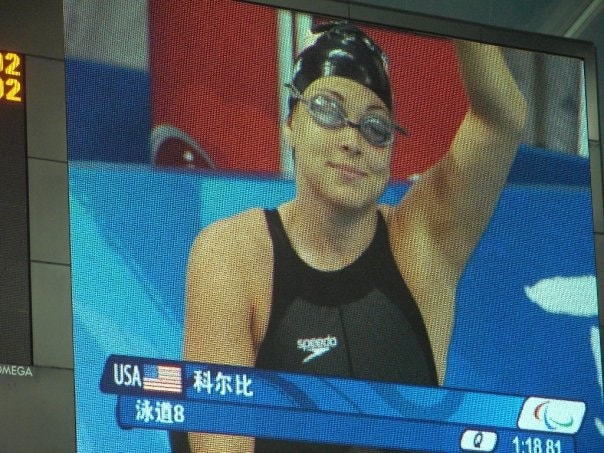Photo of Beth in China on a big screen by the starting blocks in a swim cap, goggles, and swimsuit.