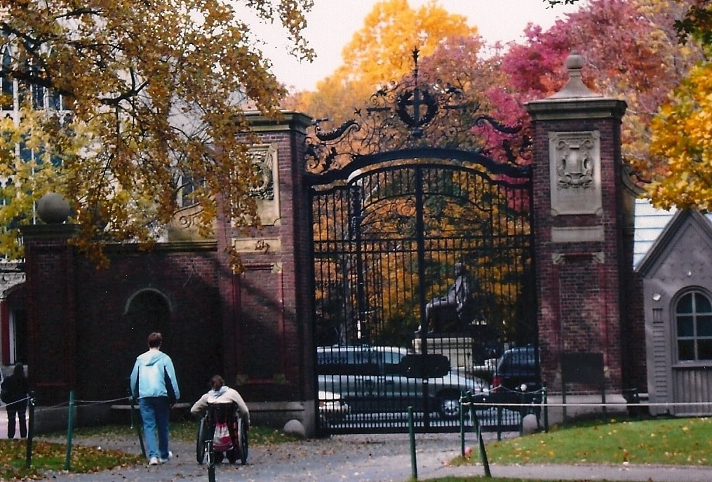 Photo of Beth pushing a wheelchair next to Cindy walking, in front of large iron and brick gates in Harvard Yard.